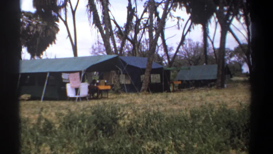 1969 Kenya 8mm frame showing safari camp tents, pale chairs, hanging fabric, dry grass, and tree trunks framing the campsite.