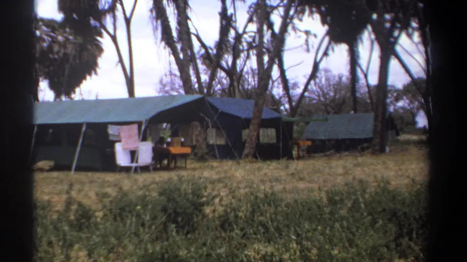 1969 Kenya campsite frame with green-topped canvas tents, folding chairs, a table, dry grass, and tall trees surrounding the camp.