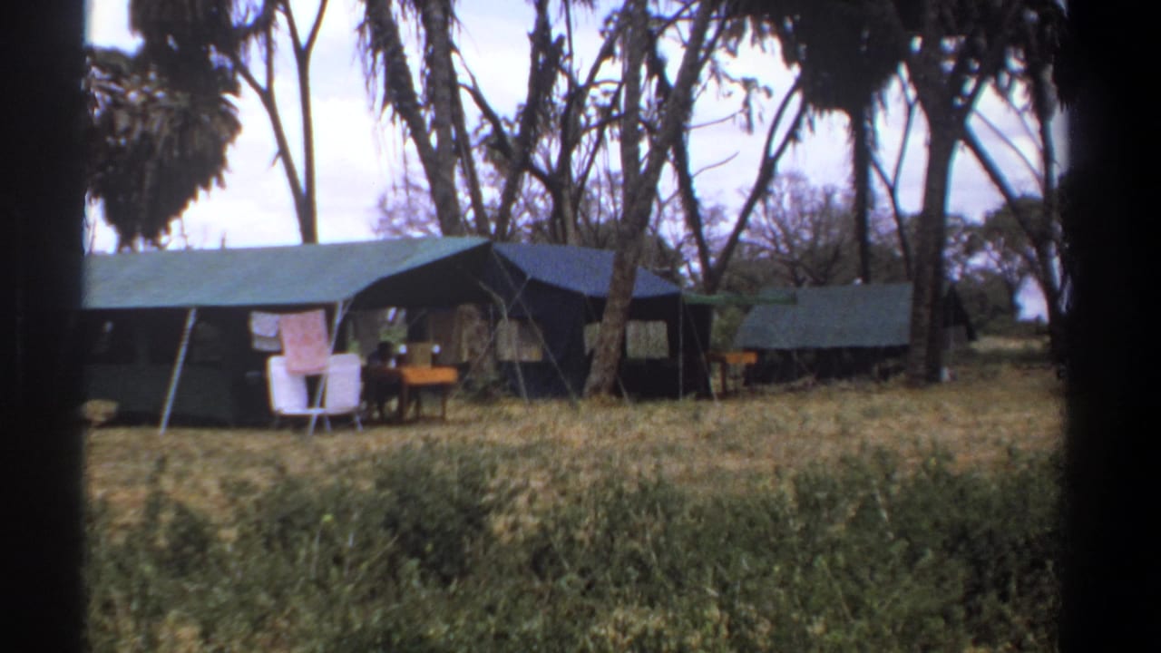 1969 Kenya home-movie frame showing canvas safari tents in dry grass with laundry hanging, white chairs, a table, and tall trees.
