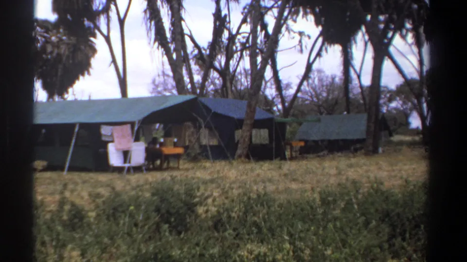 1969 Kenya home-movie frame showing canvas safari tents in dry grass with laundry hanging, white chairs, a table, and tall trees.