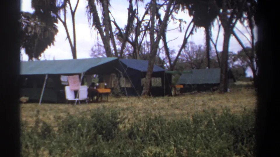 1969 Kenya frame of a quiet safari camp with canvas tents, laundry, camp chairs, table, and dry grass under tall trees.