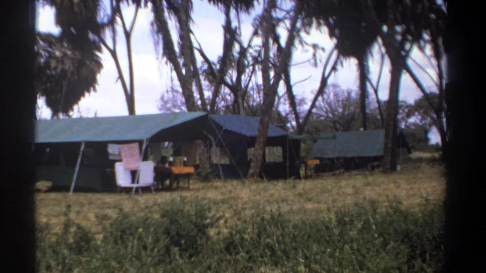 1969 Kenya home-movie still showing a tented safari camp with laundry outside, white chairs, a table, dry grass, and surrounding trees.