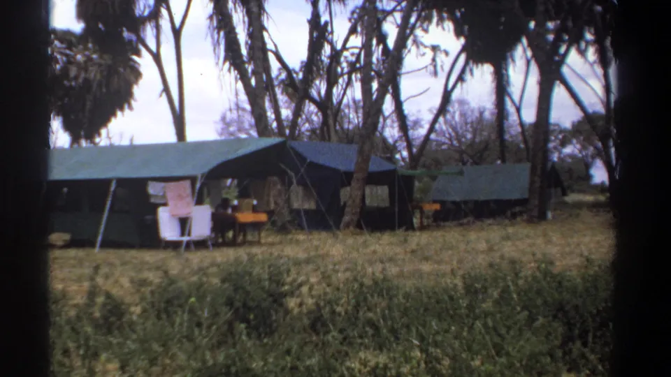 1969 Kenya safari camp frame with canvas tents, hanging laundry, chairs, a table, dry grass, trees, and a small distant person.