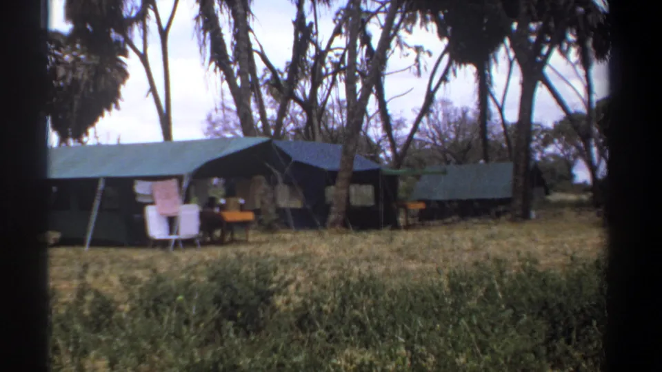 1969 Kenya safari camp with canvas tents, camp items, trees, and tall grass, framed by dark edges from the filming position.