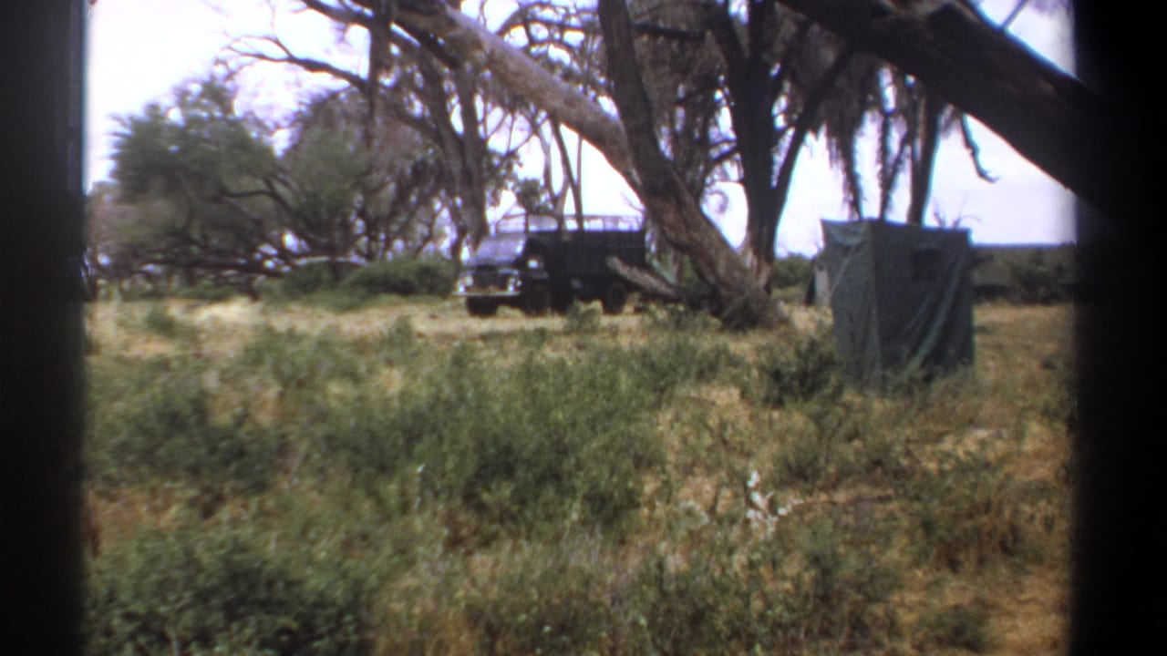 1969 Kenya safari scene showing a dark truck parked under trees near a covered canvas structure and grassland.