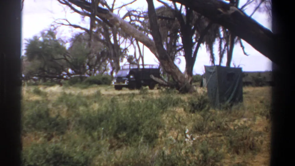 1969 Kenya home-movie view of a parked safari truck, covered camp structure, tall grass, and broad trees in savanna terrain.
