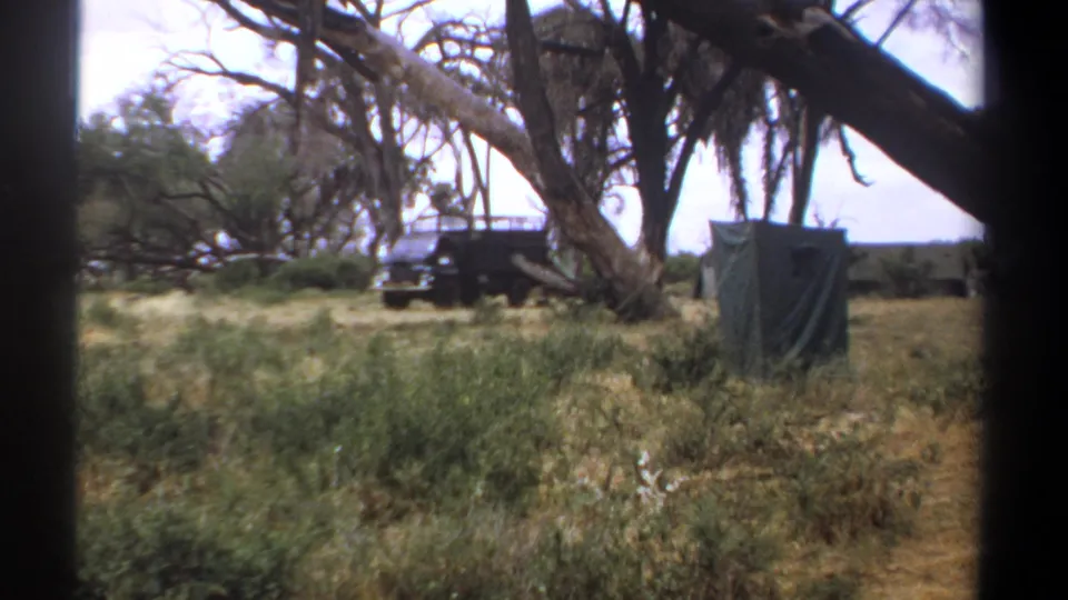 1969 Kenya safari camp view with a dark truck, canvas-covered shelter, tall grass, and trees, softened by 8mm home-movie texture.