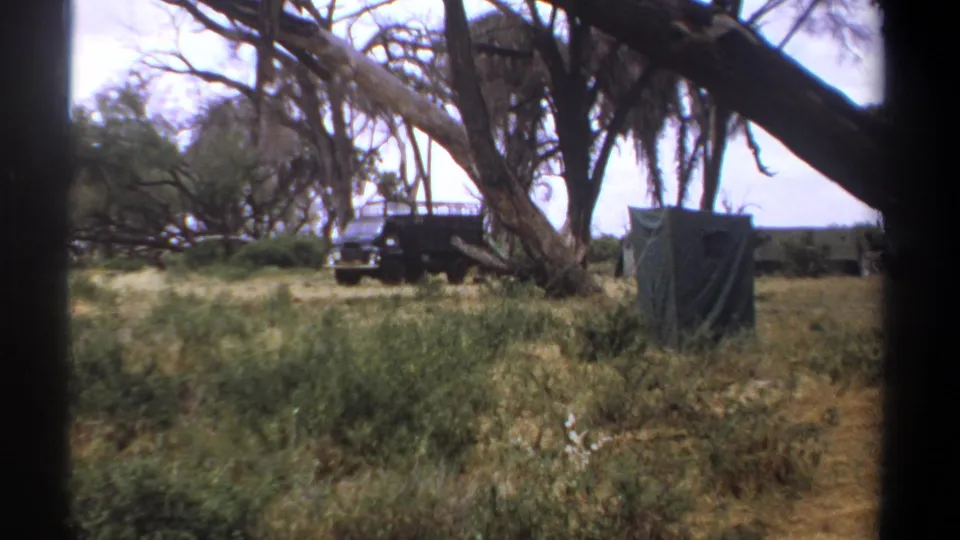 1969 Kenya home-movie frame showing a blue-gray off-road van and green canvas shelter among dry grass and trees.