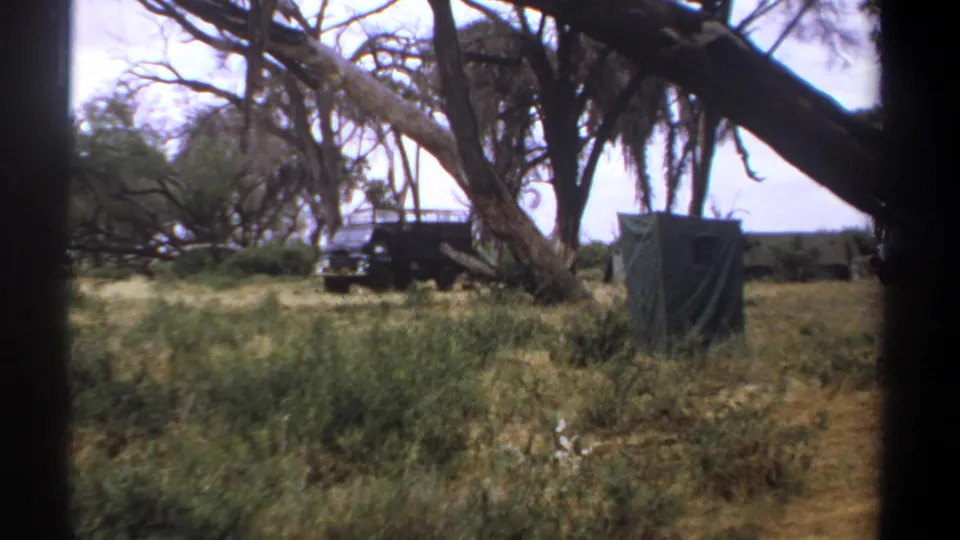 1969 Kenya footage frame with a parked off-road van, green shelter, dry grass, and dark tree trunks framing the campsite.