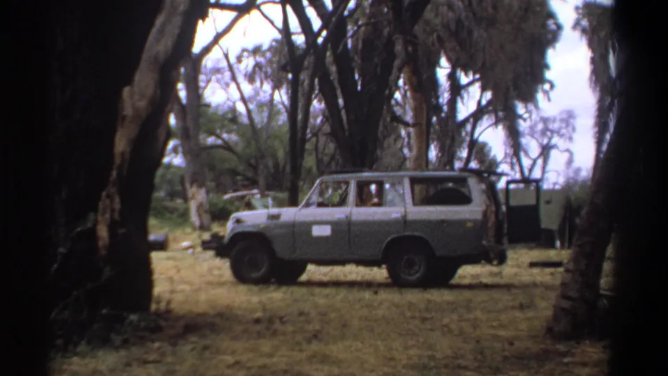 1969 Kenya home-movie frame showing the side of a blue-gray off-road van parked in a wooded dry clearing.