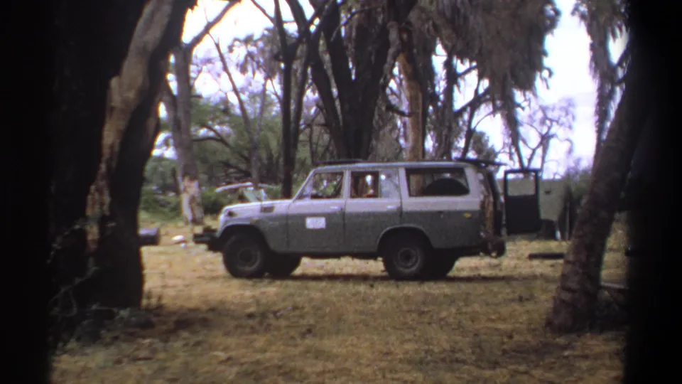 1969 Kenya frame showing a blue-gray off-road van beside a green canvas shelter in a dry wooded campsite.