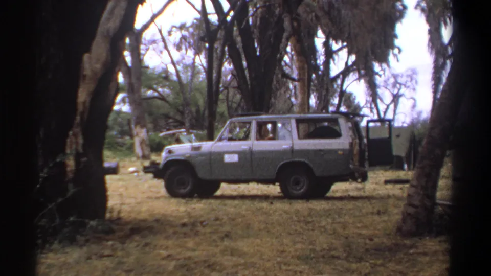 1969 Kenya 8mm frame showing a gray station wagon with a white door sign parked in a wooded clearing.