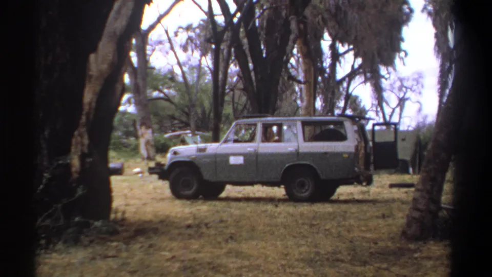 1969 Kenya travel footage frame with a gray station wagon, side sign, open rear door, dry grass, and surrounding trees.