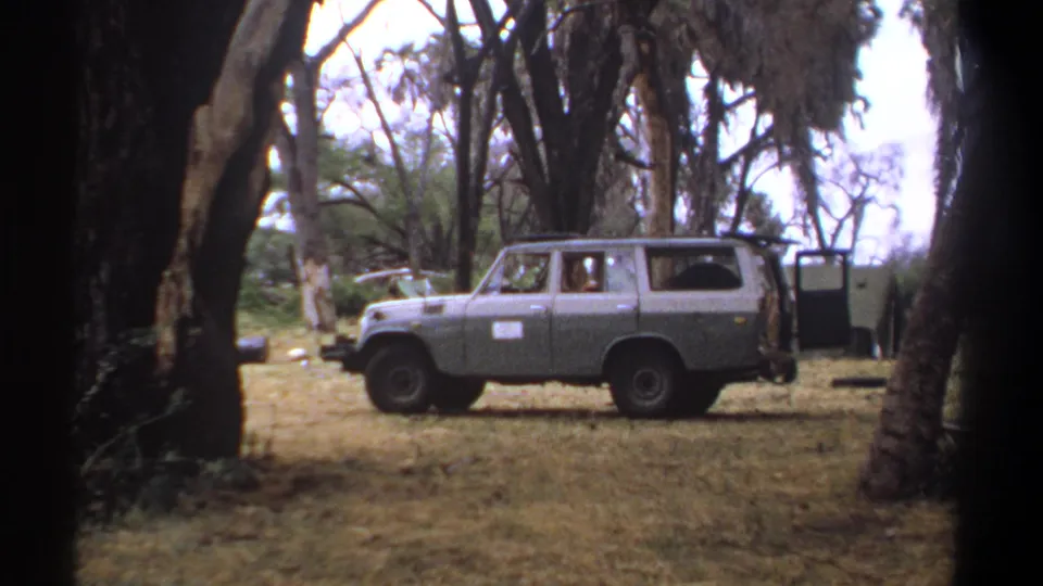 1969 Kenya 8mm frame showing a grey SUV parked beneath tall trees beside an outdoor laundry or wash area with distant people.