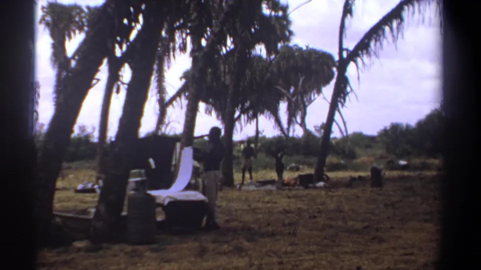 1969 Kenya home-movie frame of a person near a makeshift laundry area, with trees and several distant figures in the background.