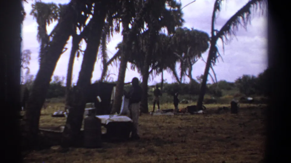 1969 Kenya 8mm frame showing an outdoor wash or laundry setup beneath tall trees, with people dispersed across the background.