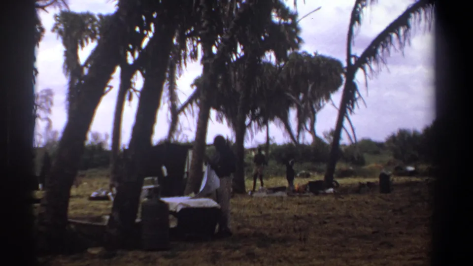 1969 Kenya home-movie frame of a shaded outdoor laundry area with tall trees, scattered items, and distant people in a rural setting.