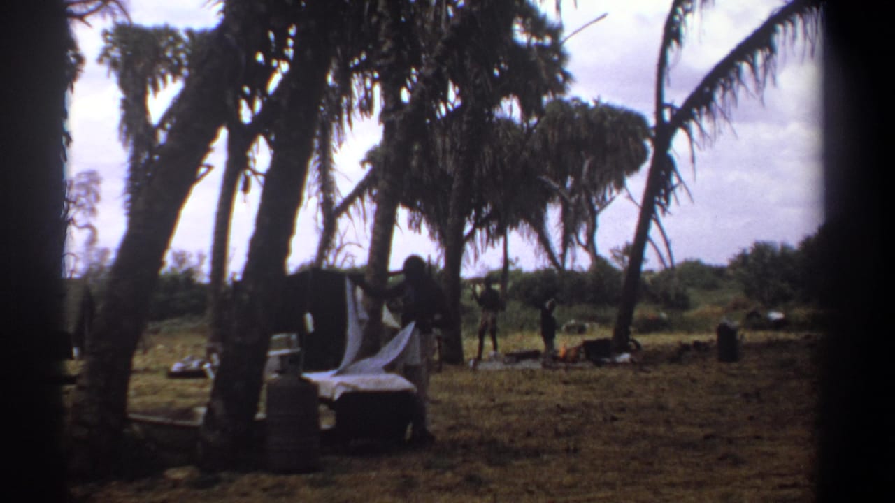 1969 Kenya 8mm frame showing a man handling white linens beneath tall trees, with outdoor furniture and distant figures in the clearing.