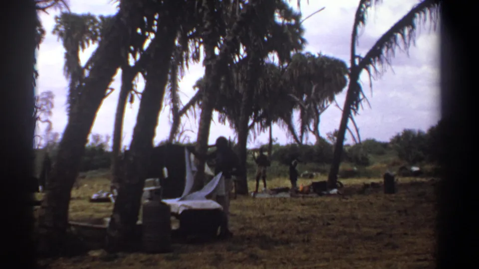 1969 Kenya 8mm frame showing a man handling white linens beneath tall trees, with outdoor furniture and distant figures in the clearing.
