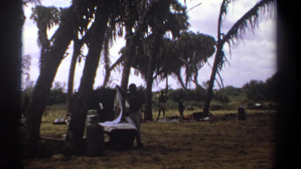 1969 Kenya home-movie frame with a man beside white linens under palm-like trees, with small figures and camp objects across the clearing.
