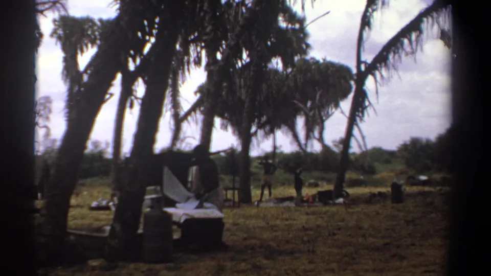 1969 Kenya 8mm frame showing a man working with a white sheet or linen beside low camp furniture in a shaded rural clearing.