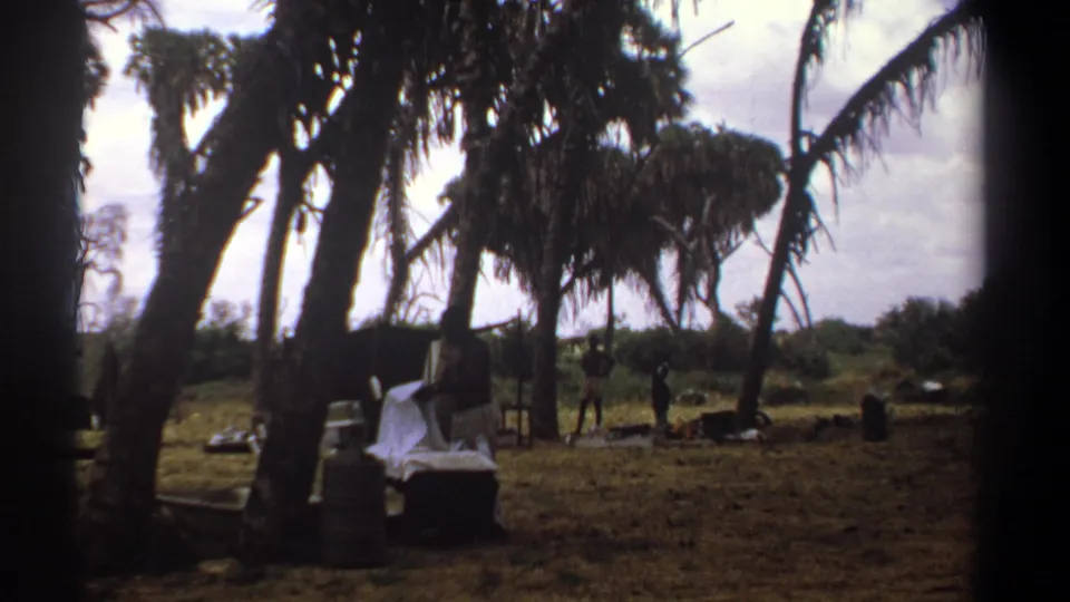 1969 Kenya home-movie frame with a man bending over white linens under tall trees, while other people and camp objects remain visible behind him.