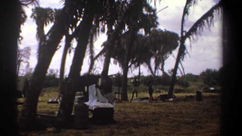1969 Kenya 8mm frame showing a rural camp under palms, with a white cloth or tarp near supplies and distant people.