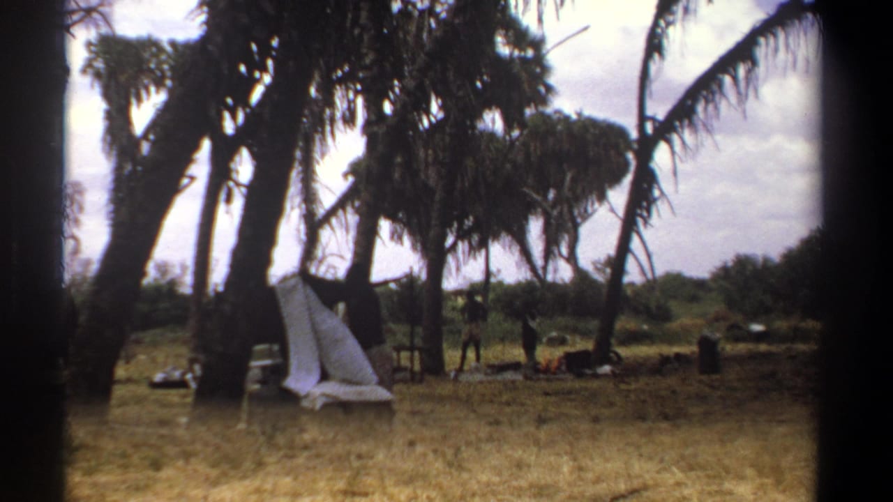 A rural Kenya camp scene from 1969, with a white cloth or tarp near palms and small distant figures among supplies.