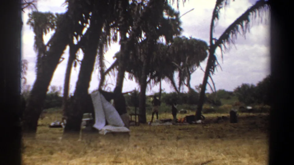 A rural Kenya camp scene from 1969, with a white cloth or tarp near palms and small distant figures among supplies.