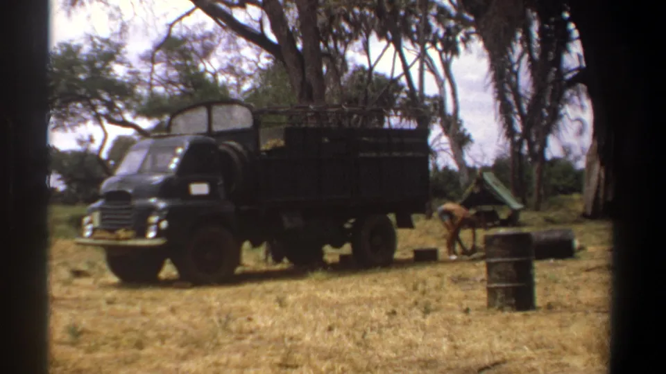 1969 Kenya 8mm frame with a black work truck parked in a dry camp area as a person bends near barrels and supplies.
