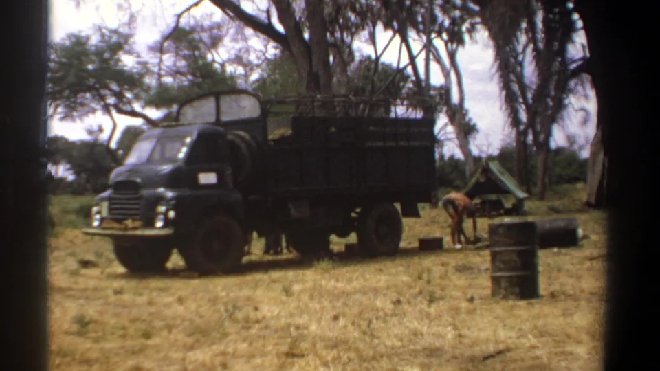 1969 Kenya home-movie frame showing a dark dump truck at a dry campsite with trees, drums, a small tent, and a worker bending nearby.