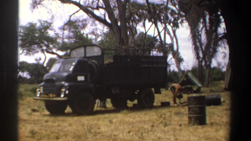 1969 Kenya frame showing a dark dump truck, scattered campsite equipment, oil drums, and a worker bending near a small green tent.