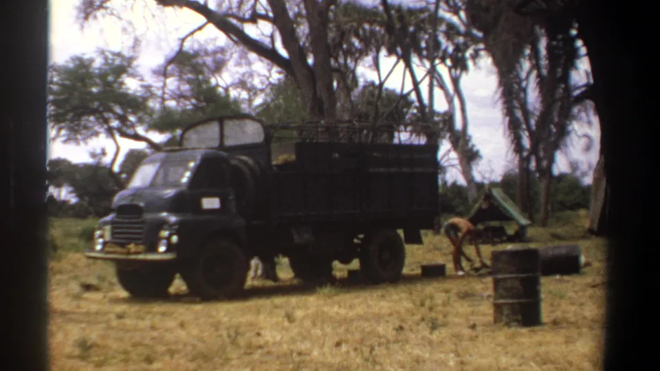 1969 Kenya 8mm frame showing a dark green military truck under trees with barrels, crates, and camp gear nearby.