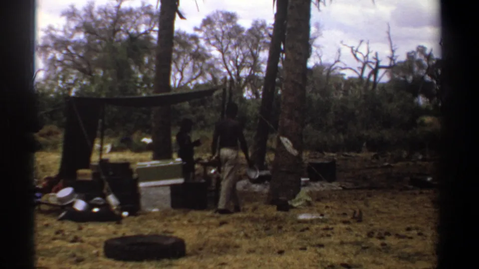 1969 Kenya 8mm frame showing uniformed figures near a makeshift shelter, crates, a tire, and scattered field equipment.