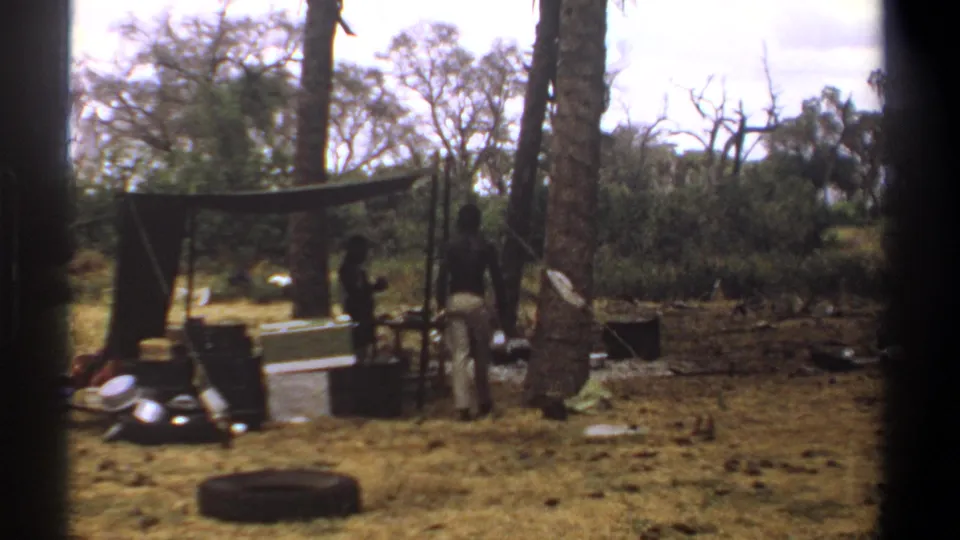 1969 Kenya 8mm frame showing two men near a makeshift tarp shelter, containers, cookware, and a tire in a dry forest campsite.