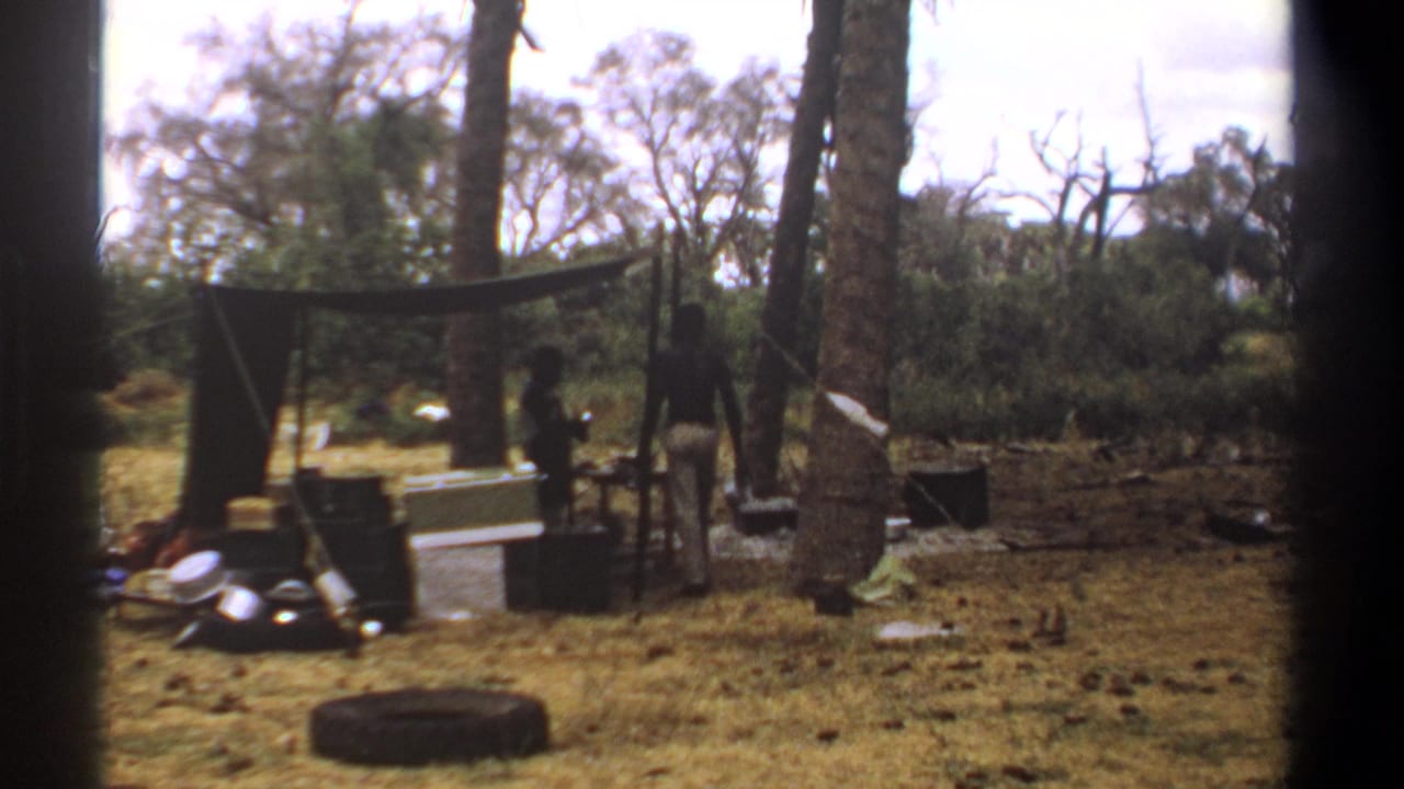 1969 Kenya home-movie frame with two figures partly visible around a tarp shelter, stacked supplies, trees, and dry grass.