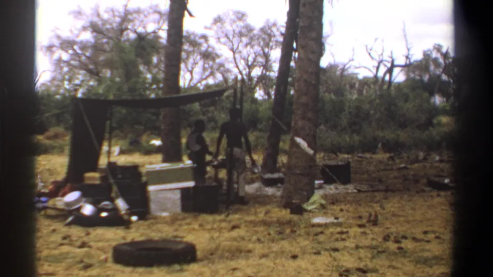 1969 Kenya 8mm frame showing two men beside a tarp shelter with crates, cookware, a tire, dry grass, and trees around the campsite.