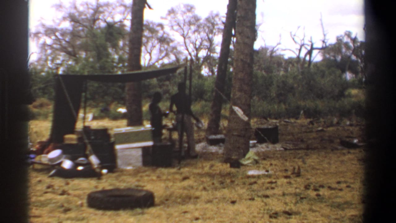 1969 Kenya home-movie frame with two dark figures near a pole-supported tarp shelter amid dry grass and wooded brush.