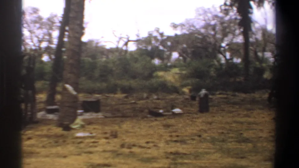 1969 Kenya 8mm frame looking across a dry campsite with trees, scattered gear, a tarp edge, and a distant standing figure.