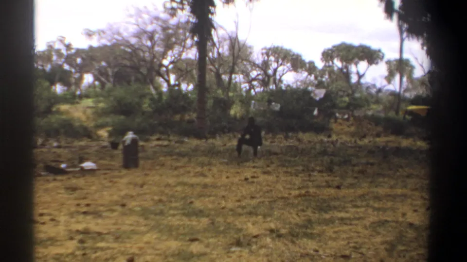 1969 Kenya home-movie frame showing a wide dry clearing with a silhouetted person, tall trees, and scattered camp items.