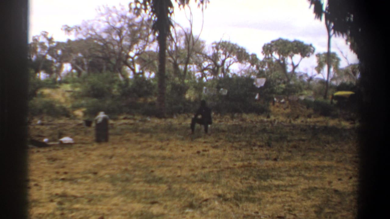 1969 Kenya 8mm frame showing a dry campsite clearing with two distant people, scattered trees, and dark vertical edge obstruction.