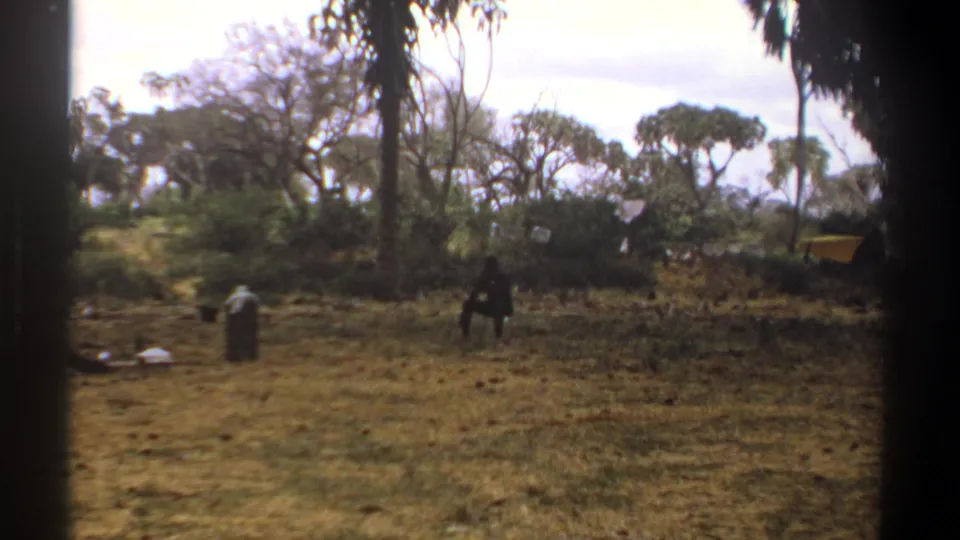 1969 Kenya 8mm frame showing distant figures in a dry clearing with trees, scattered gear, and soft home-movie exposure.