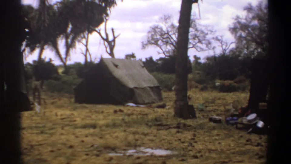 1969 Kenya home-movie frame showing a canvas tent in a dry wooded clearing with scattered camp items and tree trunks.