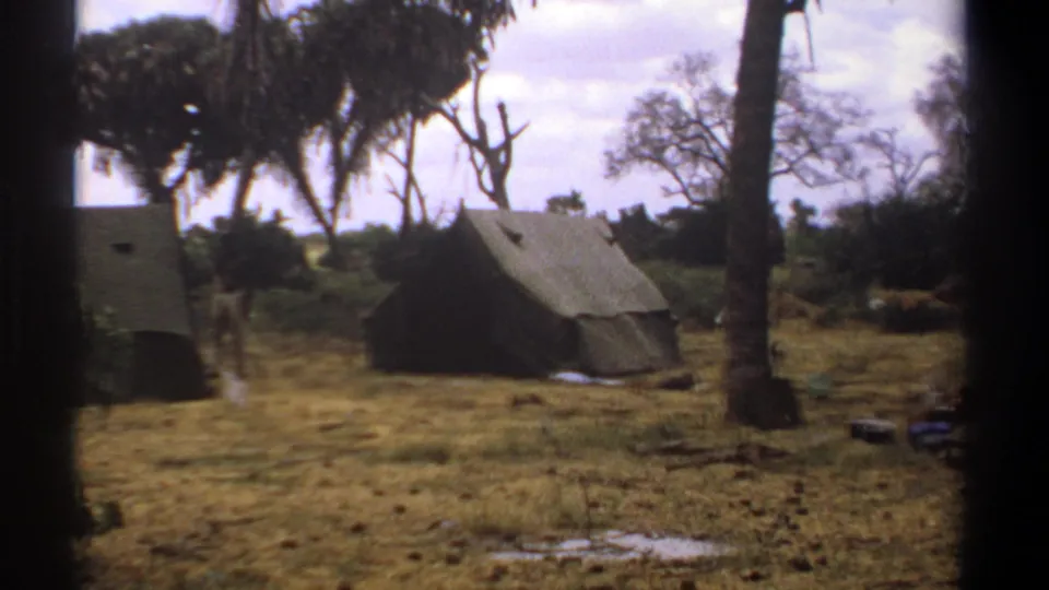 1969 Kenya 8mm frame showing two canvas tents in a dry tropical camp, with palm trees, shrubs, cloudy sky, and a distant person near the tents.