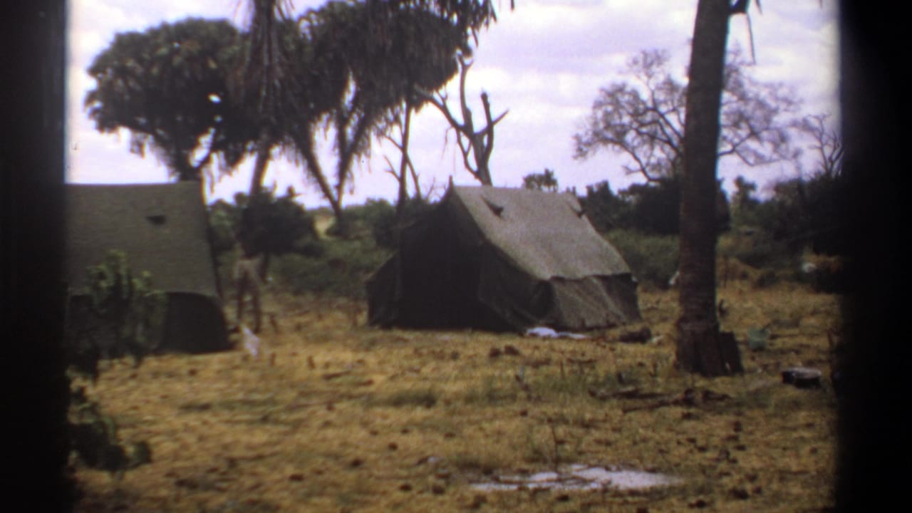 1969 Kenya 8mm frame of a sparse tented camp, with two canvas tents, palm trunks, dry grass, and a small figure moving between them.