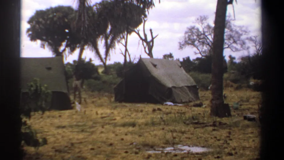 1969 Kenya 8mm frame of a sparse tented camp, with two canvas tents, palm trunks, dry grass, and a small figure moving between them.