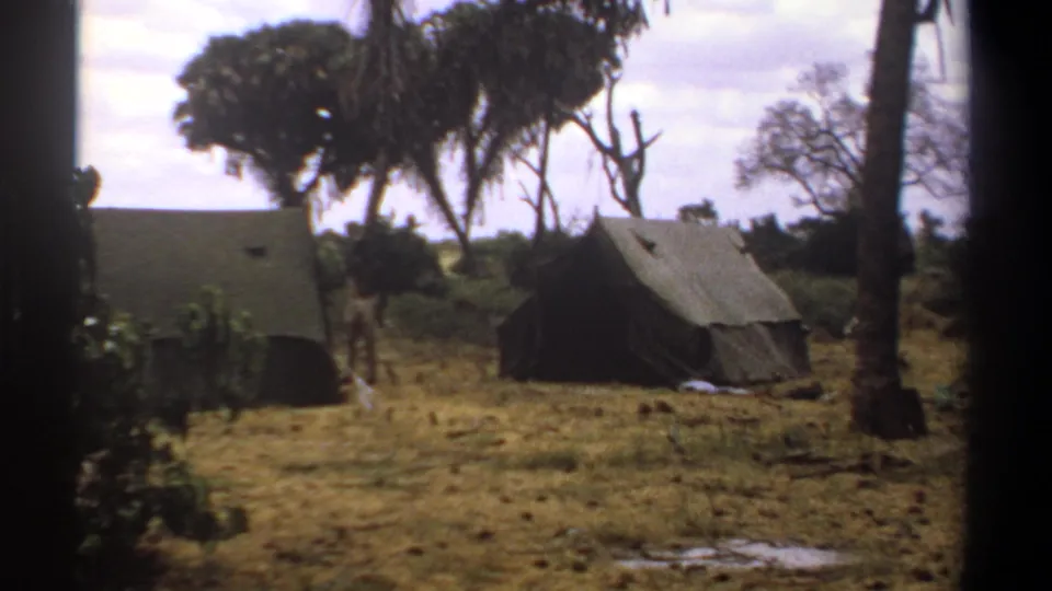 1969 Kenya 8mm frame showing canvas tents across dry grass, partly framed by dark film edges and nearby foliage in a tropical camp.