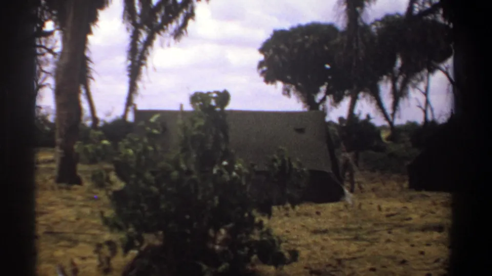 1969 Kenya 8mm frame showing a dry campsite with palms, shrubs, and canvas tents in the distance.
