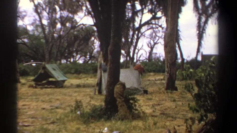 1969 Kenya 8mm frame showing a green A-frame tent, pale canvas shelter, palms, and a distant person at camp.