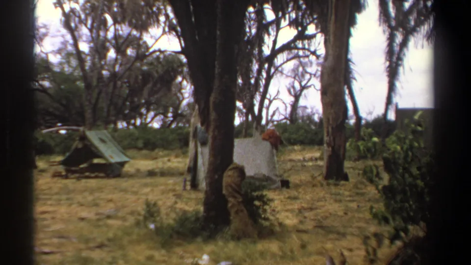 1969 Kenya 8mm frame of the campsite clearing with a green tent, pale shelter, palms, and a distant person.
